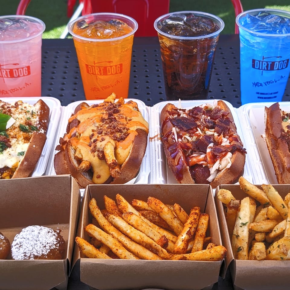 Dirt Dog loaded hot dogs with fries, powdered pastries, and four colorful iced drinks arranged on a patio table.