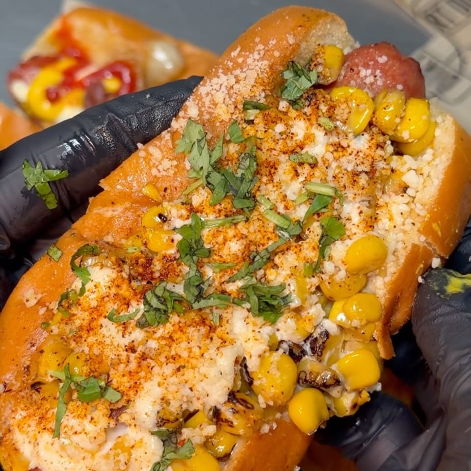 Close-up of a hot dog topped with corn, cheese, cilantro, and chili powder, held by gloved hands.