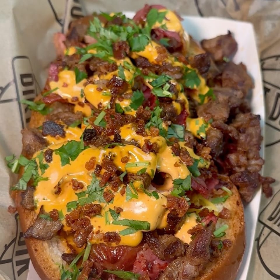Two gourmet hot dogs topped with steak, cheese sauce, bacon bits, and cilantro in paper-lined trays.