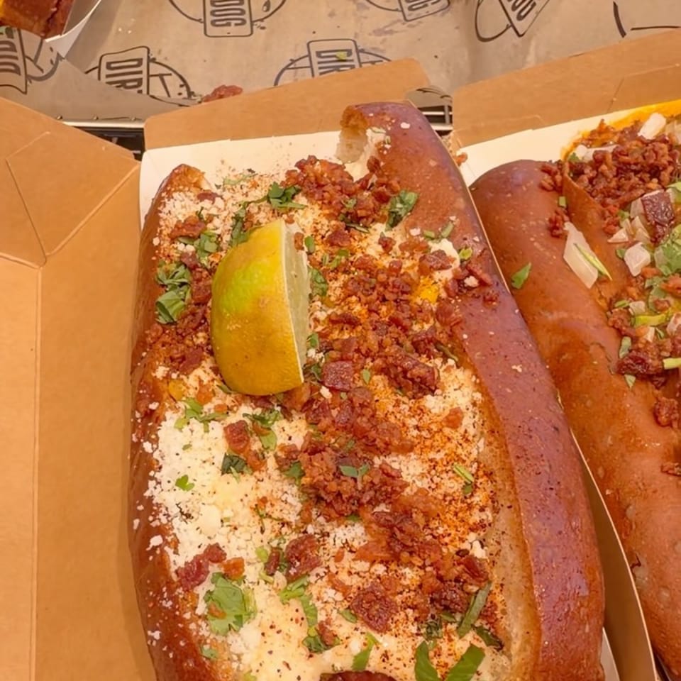 A Dirt Dog hot dog in a pretzel bun, topped with bacon, cheese, cilantro, and a lime wedge.