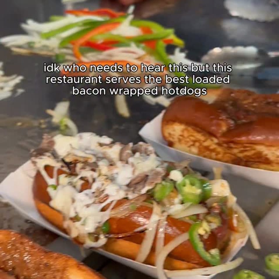 Three loaded hotdogs with various toppings, including bacon, onions, and peppers, on a metal surface.