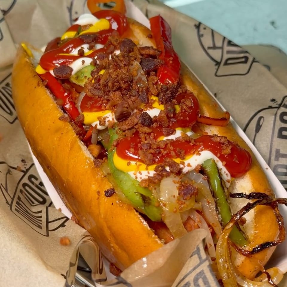 A loaded hot dog with bacon, peppers, onions, avocado, ketchup, and mustard in a paper-lined basket.