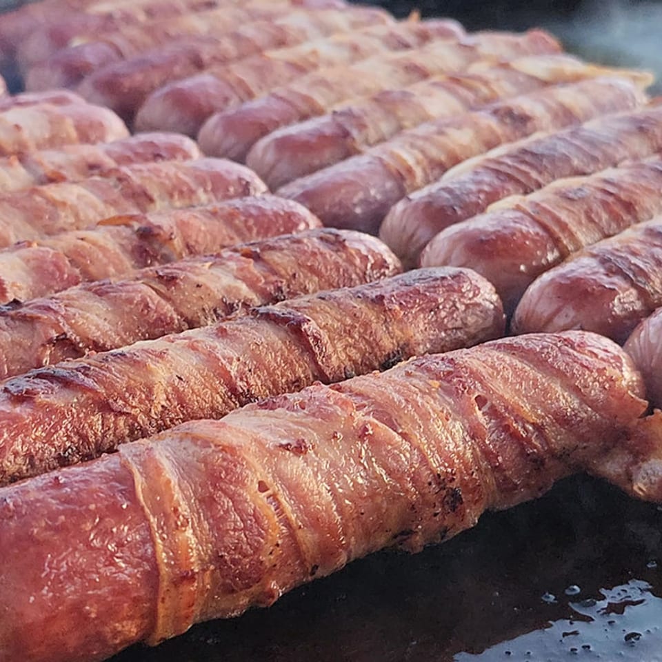 Rows of bacon-wrapped sausages sizzle on a flat-top grill, glistening with cooking juices.