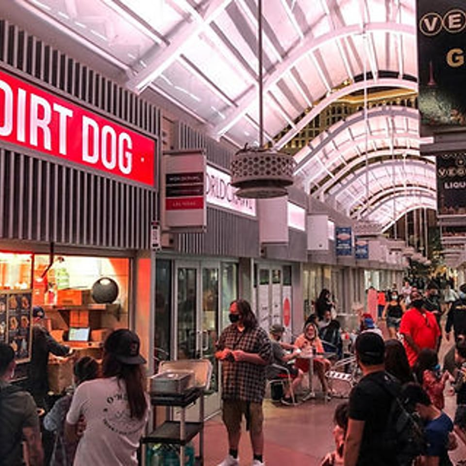 A bustling indoor market with people walking past the Dirt Dog restaurant and other shops.
