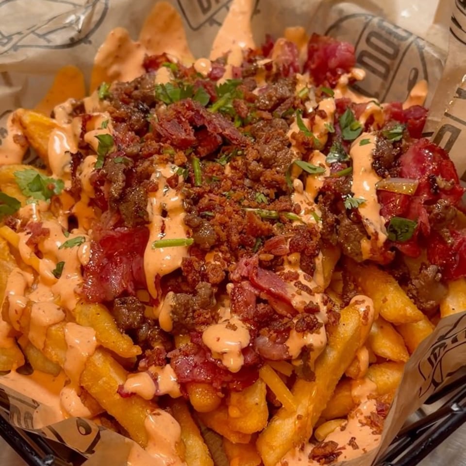 Loaded fries with crumbled bacon, meat, sauce, and parsley in a branded basket.