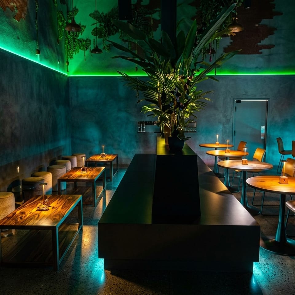 Moody modern restaurant interior with teal lighting, tropical plants, wooden tables, round seating, and candlelit tables.