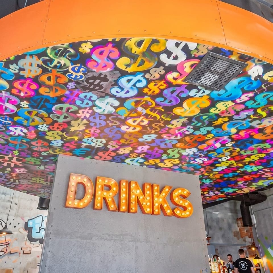 Restaurant interior featuring a colorful dollar-sign mural ceiling and illuminated orange “DRINKS” sign.