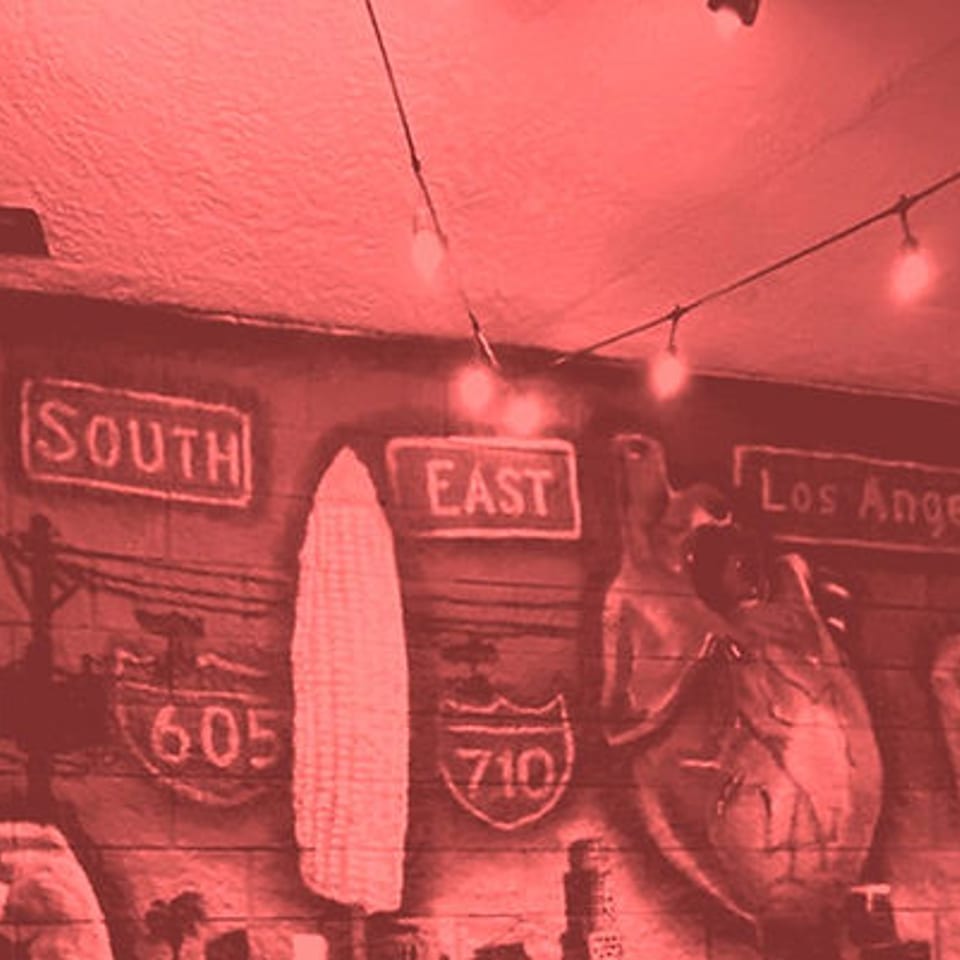 Red-tinted mural with "South East Los Angeles" and freeway signs, illuminated by string lights.
